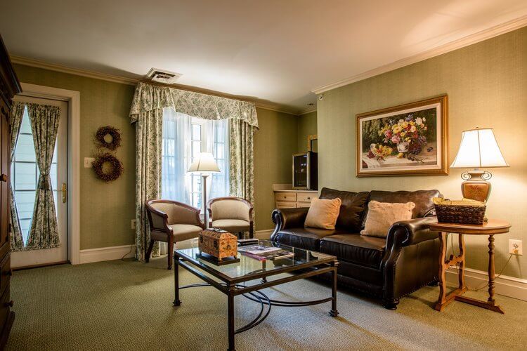 A cozy living room with green carpet and wallpaper. There's a black leather sofa with beige cushions, a glass coffee table, and two white armchairs with wooden frames. Two lamps provide warm lighting. A painting of flowers hangs on the wall, near a window with floral curtains.