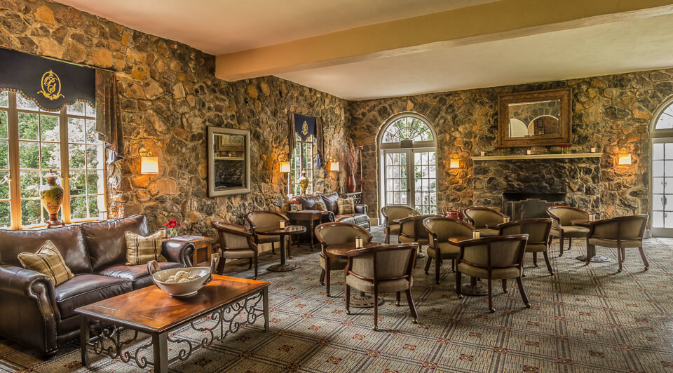 A cozy, rustic living room with stone walls, a large rug, and several seating areas. There are leather couches, wooden chairs with cushions, and small tables. Two arched windows and a door let in natural light. A fireplace is flanked by wall sconces and artwork.