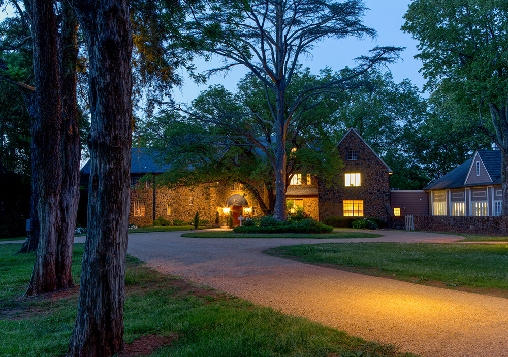 A large stone house with lit windows stands surrounded by trees at dusk. A curved gravel driveway leads up to the front entrance, illuminated by warm outdoor lighting. The scene is peaceful with a mix of natural and artificial light.