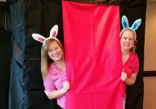 Two women wearing bunny ear headbands stand beside a red curtain at a photobooth. Both are smiling and dressed in pink shirts. The setup is surrounded by a black backdrop, providing contrast to the bright red curtain and their cheerful appearance. Image