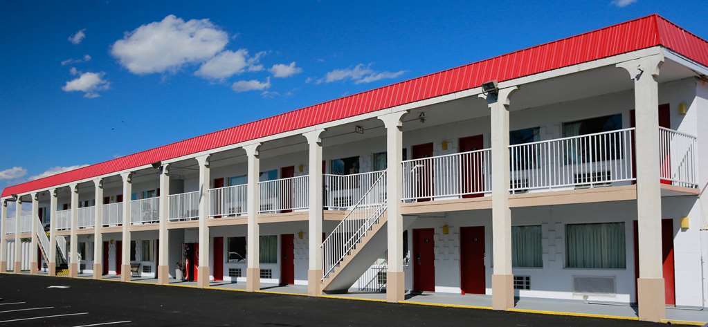 A two-story motel with a red roof and white exterior. The building has exterior corridors with railings and several doors leading to individual rooms. A bright blue sky with a few clouds is visible above. The parking lot in front of the motel appears empty.