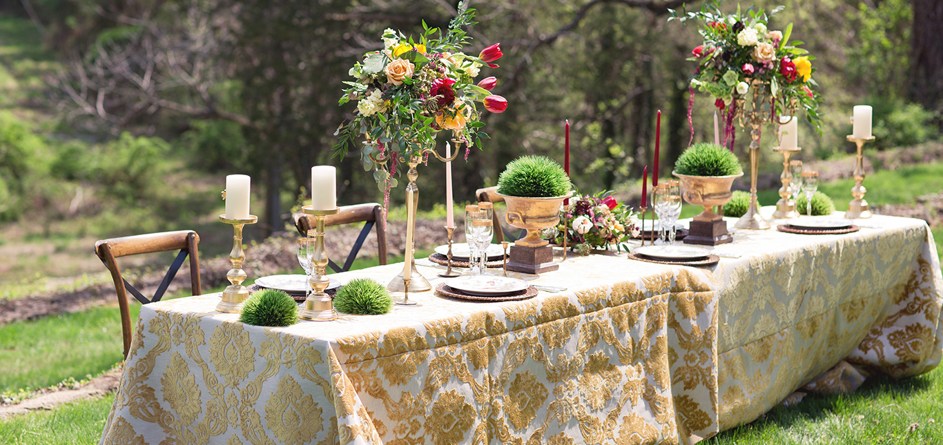 A beautifully set outdoor dining table with a patterned gold and white tablecloth, adorned with elegant floral centerpieces and candle holders. Plate settings are arranged neatly, and greenery adds a touch of natural decor. The background features lush greenery.