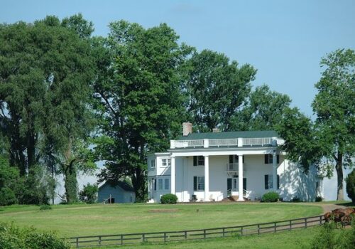 A large white colonial-style house with green shutters and a green roof stands amidst lush greenery. Tall trees surround the house, and a white fence lines the foreground, enclosing a well-maintained lawn under a clear blue sky. Image