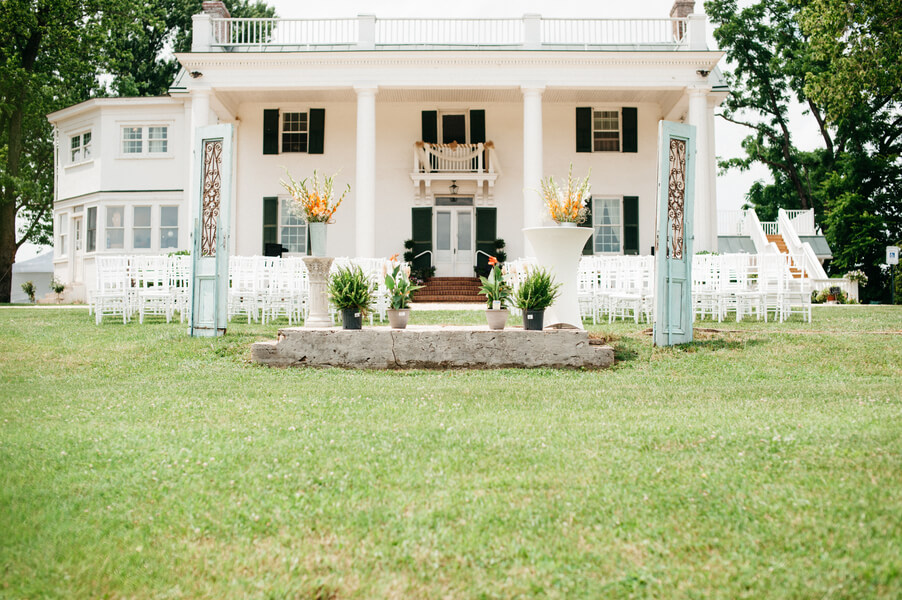 A white two-story building with a large porch and green shutters is set up for an outdoor event. Numerous white chairs are arranged in rows on a grassy lawn, facing the building. Two tall flower arrangements flank the walkway leading to the building.
