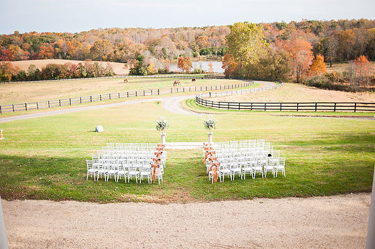 An outdoor wedding ceremony setup featuring rows of white chairs arranged on a grassy field, facing an arch adorned with flowers. In the background, there's a winding path, wooden fences, and a picturesque landscape of trees with autumn foliage.