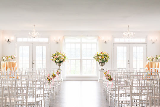 A bright, elegant wedding ceremony setup with rows of white chairs lined up facing a large window. The space is decorated with lush floral arrangements of pink and white flowers, some on the floor and others in tall vases. Chandelier lighting adds a touch of sophistication.