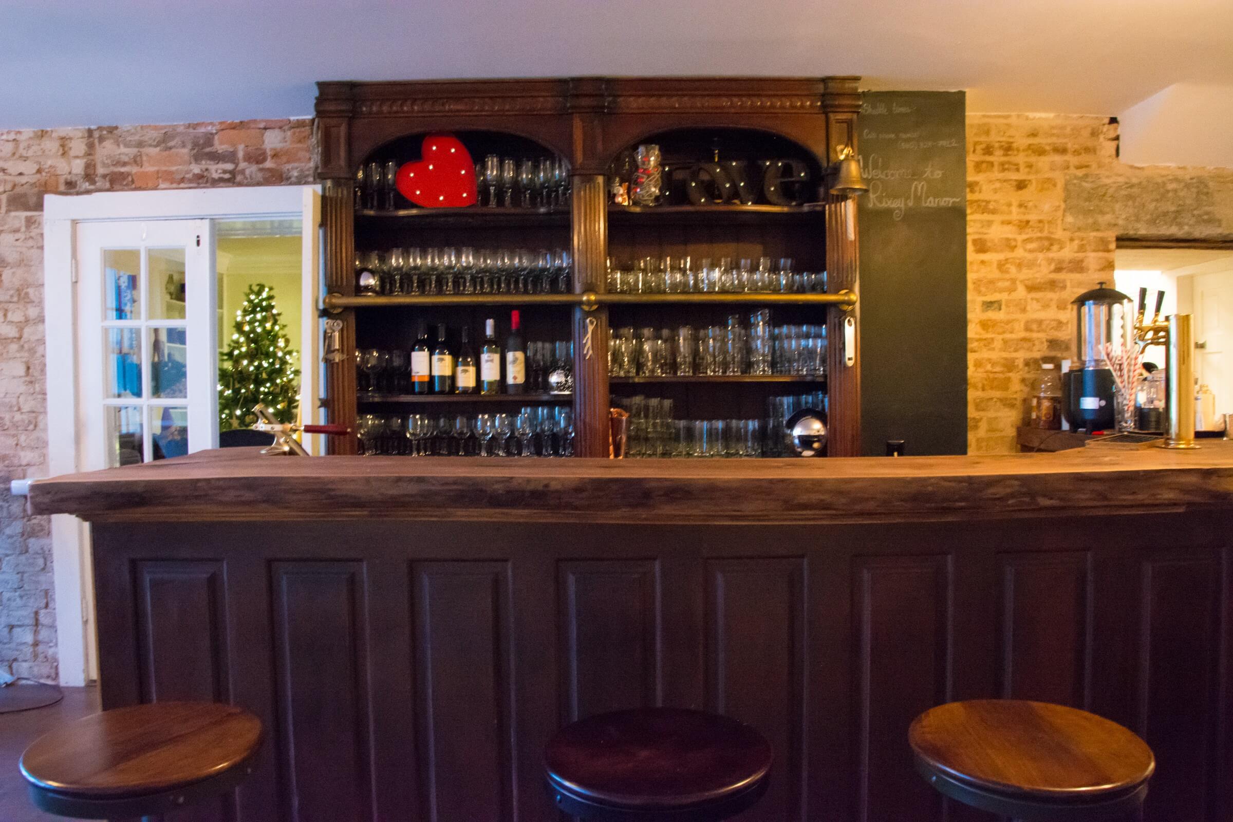 A cozy bar with a wooden counter and three stools in front. Behind the counter, shelves are stocked with glasses and assorted bottles. Decorations include a heart-shaped ornament and a small Christmas tree in the background near a white-framed door.