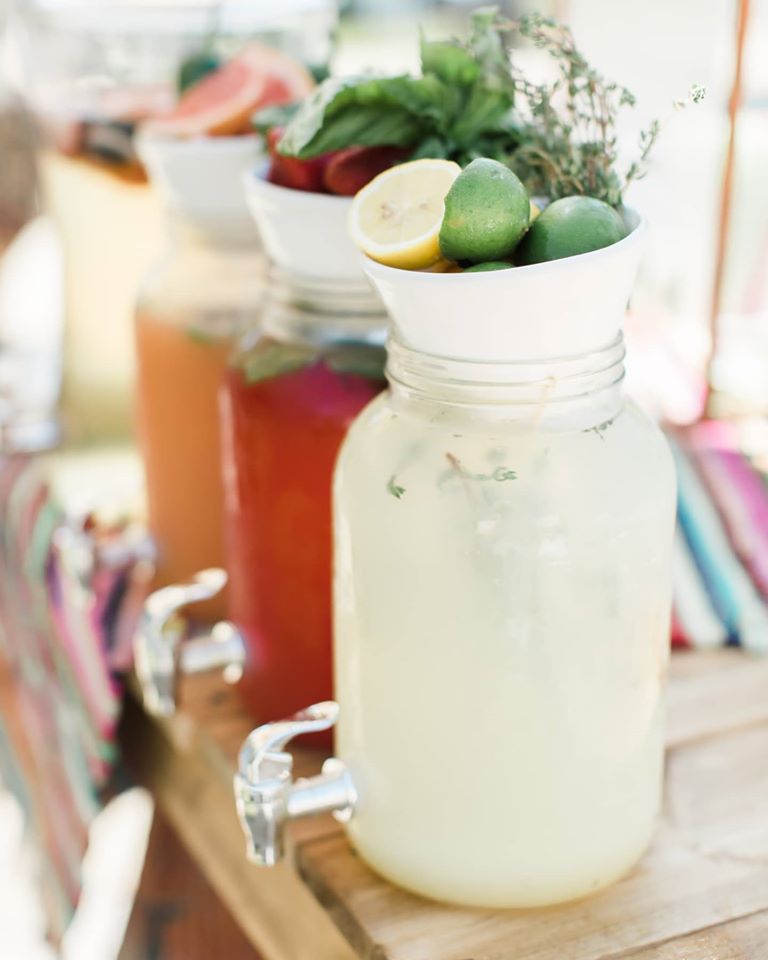 Three glass beverage dispensers filled with different colorful drinks are on a table. The front dispenser contains lemonade garnished with lemons, limes, and herbs. The middle and rear dispensers hold fruity drinks and are similarly garnished with fresh fruits and herbs.