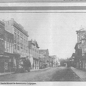 Black and white historical photo of Davis Street in downtown Culpeper. The street is lined with old buildings, some with visible signs for various businesses. The scene is quiet, with no visible cars or people. A printed caption at the bottom reads, 