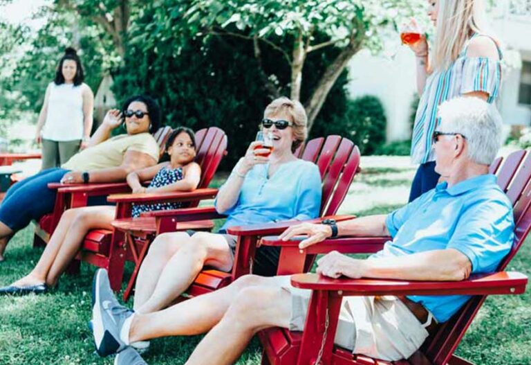A group of people relaxes on red Adirondack chairs in a lush, grassy yard. They are chatting and enjoying drinks. Some are smiling, and a woman stands on the right, holding a drink. Trees and a building are visible in the background.