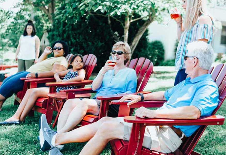 A group of people relaxes on red Adirondack chairs in a lush, grassy yard. They are chatting and enjoying drinks. Some are smiling, and a woman stands on the right, holding a drink. Trees and a building are visible in the background.