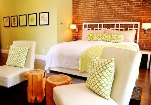A modern bedroom with a white bed frame, green and white bedding, and matching accent pillows. The room has a brick accent wall, five nature-themed framed pictures, two cream-colored chairs with patterned cushions, and two wooden stump side tables. Image