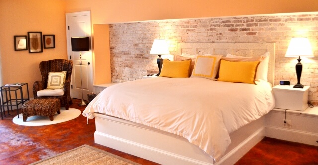 A cozy bedroom with a large white bed adorned with yellow pillows, flanked by two bedside tables with lamps. A wicker chair with a cushion and a small side table stand in the corner. The room features a white brick accent wall and warm lighting.