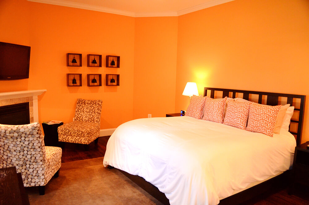 A cozy bedroom features a large bed with a white comforter and patterned pillows. The walls are painted a warm orange, and the room includes a bedside lamp, two patterned chairs, a wall-mounted TV, and framed artwork above the fireplace.