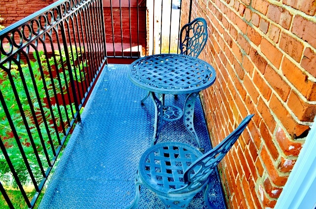A small wrought iron balcony with two matching chairs and a round table, both featuring intricate lattice designs. The balcony is adjacent to a red brick wall and overlooks greenery below. The scene is bright and inviting, suggesting a sunny day.