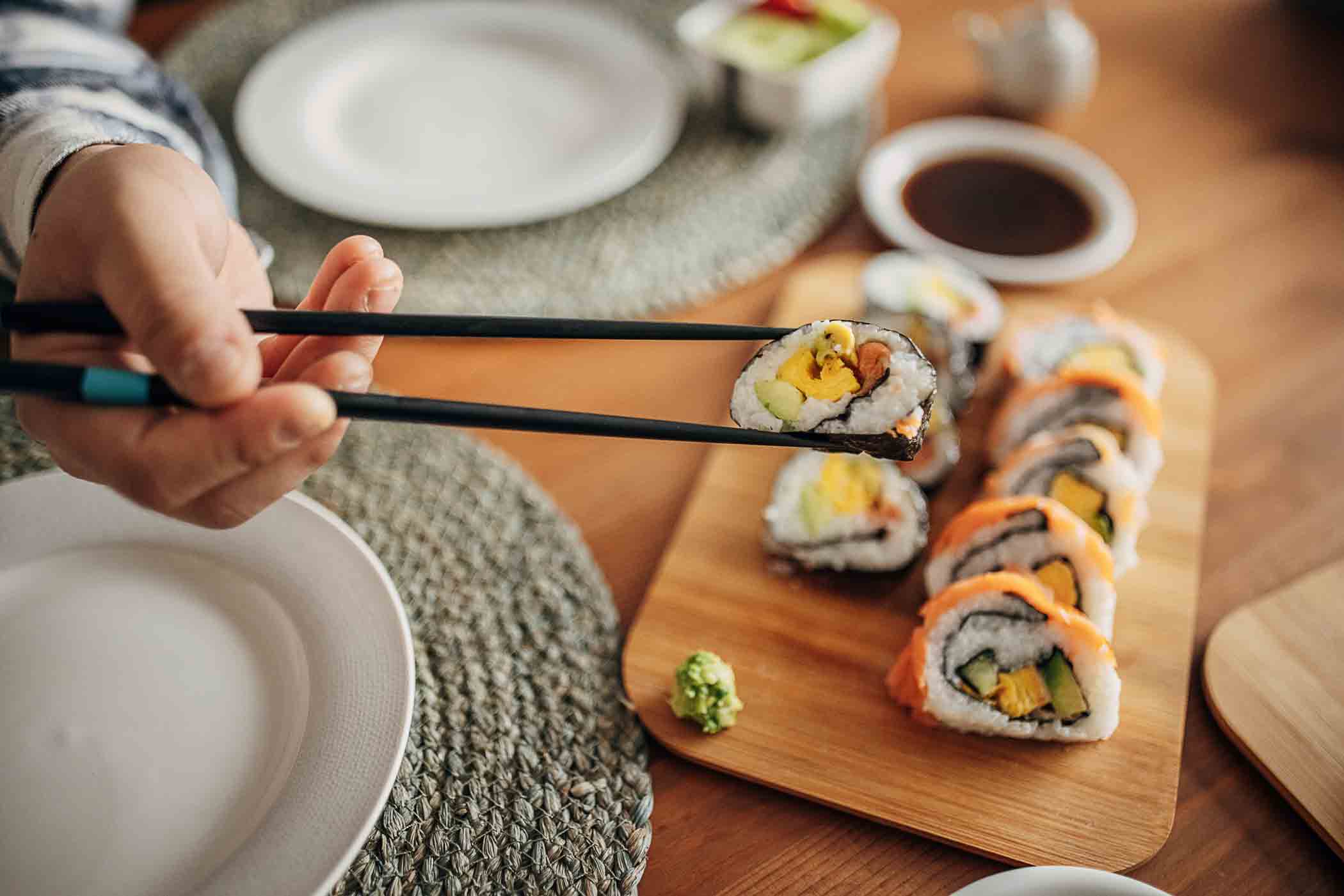 A person uses chopsticks to hold a piece of sushi over a wooden board filled with various sushi rolls. The table is set with plates, and a small dish of soy sauce. A small green wasabi mound is present on the board.