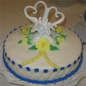 A round white cake with elegant blue and yellow decorations. The cake features purple icing details along the bottom edge and yellow roses on top. At the center of the cake, there is a decorative topper with intertwined hearts and two white doves. Image