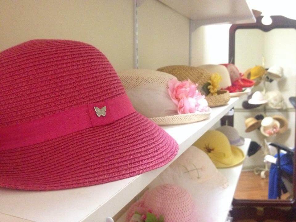 A display of various hats on white shelves. In the foreground, a bright pink hat with a butterfly pin is prominent. Behind it, there are hats in different colors and styles, some adorned with flowers and other decorative items. The background shows more hats and a mirror.