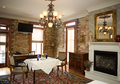 A cozy dining area features a table set for two with a white tablecloth and blue and clear glassware. The room has exposed brick walls, a fireplace with a white mantle, a decorative mirror, and a chandelier. There is a small TV mounted on the wall, wooden flooring, and a colorful rug. Image