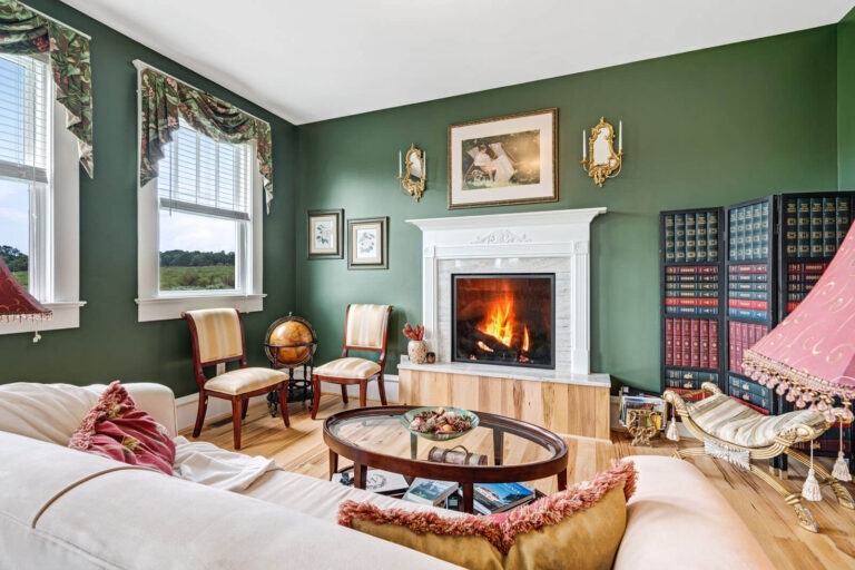 A cozy living room with green walls features a lit fireplace with a white mantle. Two chairs sit beside a globe, while a bookcase stands in the corner. A sofa with cushions, a round coffee table, and windows with ornate curtains add to the inviting atmosphere.