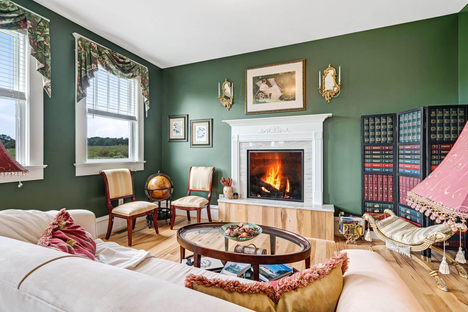 A cozy living room with green walls features a lit fireplace with a white mantle. Two chairs sit beside a globe, while a bookcase stands in the corner. A sofa with cushions, a round coffee table, and windows with ornate curtains add to the inviting atmosphere.