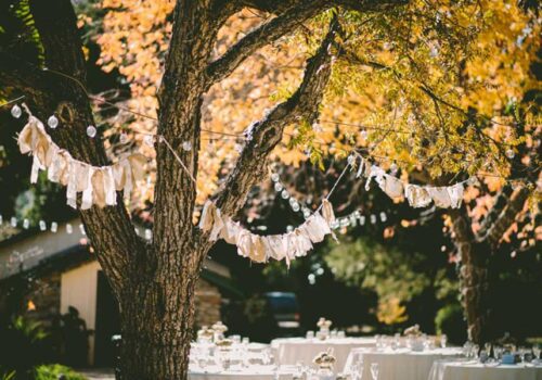 A rustic destination wedding setup with tables and chairs arranged under trees adorned with string lights and garlands. The leaves on the trees have turned golden, creating an inviting and warm autumn atmosphere.