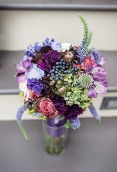 A colorful floral bouquet in a glass vase featuring a mix of flowers including purple, blue, green, and pink blooms, accented with small berries and greenery, placed on steps. Image