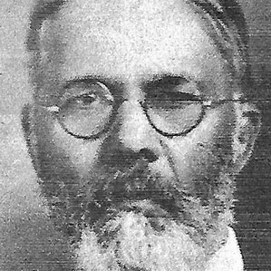 Black and white photograph of an older man with round glasses, a full beard, and short hair. He is facing the camera directly, and the background is plain and indistinct.