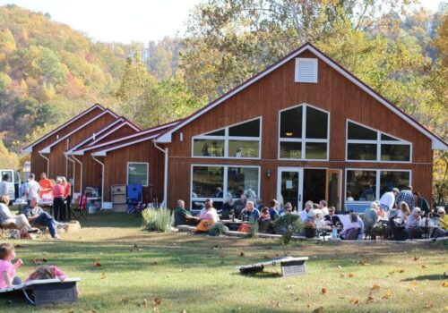 A group of people gathered outside a wooden building with large windows, enjoying a sunny day. Some people are sitting at picnic tables, while others are playing games on the grass. The surrounding area is filled with autumn-colored trees and hills. Image