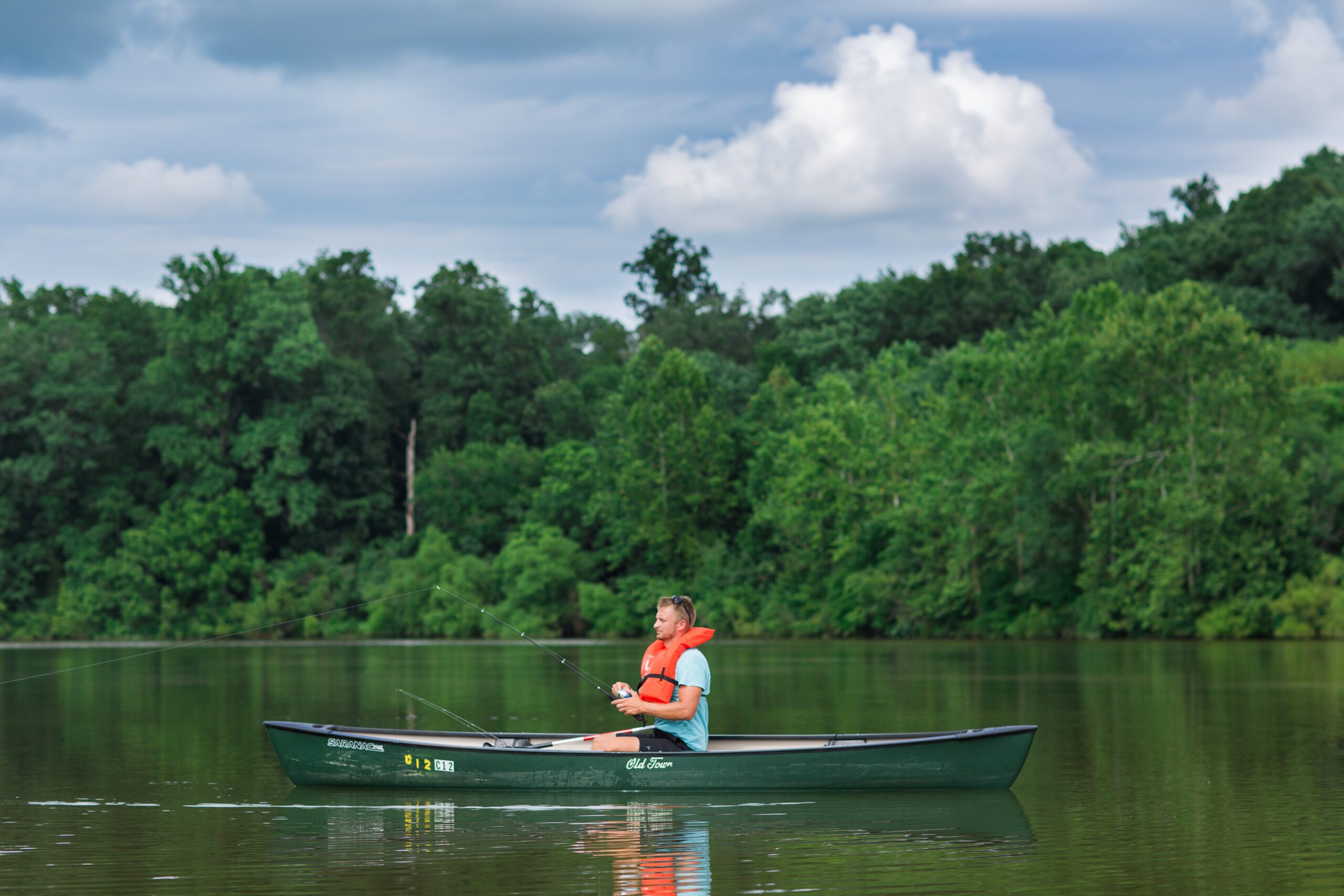 A person wearing a life jacket sits in a green canoe on a calm lake, holding a fishing rod. The surrounding area is lush with dense green trees, and the sky is partly cloudy.