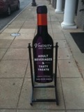 A sidewalk sign resembling a bottle of wine stands on a concrete walkway outside a building. The sign advertises 