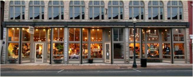 A wide-angle view of a large storefront with multiple large windows displaying a well-lit interior. The building has a classic, historical design with a row of small upper windows above the main entrance. The street in front is empty with a single lamppost visible.