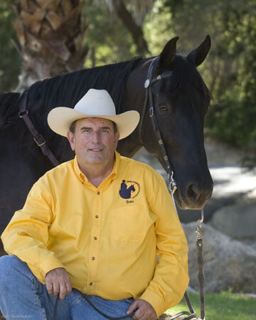 A man in a yellow shirt and white cowboy hat sits next to a black horse. They are outdoors with trees and rocks in the background. The man appears calm and content, holding the horse's reins casually.
