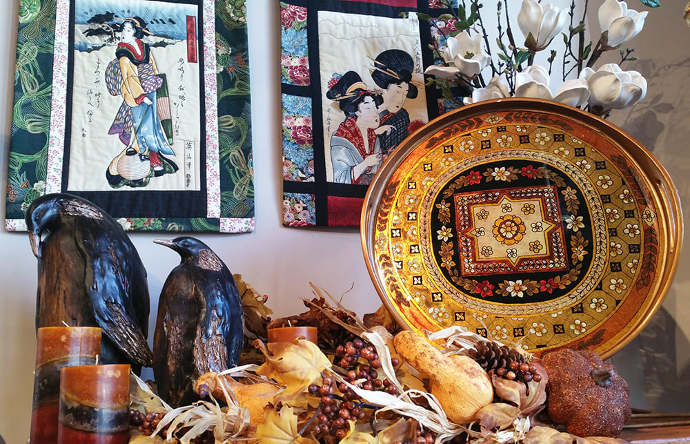 A vibrant display features a large ornate plate with intricate patterns, two bird sculptures, and a variety of decorative items including dried fruits and candles. Behind the arrangement are two colorful textile artworks depicting traditional Japanese figures and scenes.