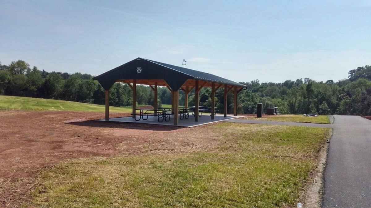 A covered pavilion with picnic tables and a concrete floor is situated in a park. The pavilion is open on the sides and has a gable roof. Surrounding it are a grassy field, a path, and tree-lined areas in the background under a clear, blue sky.