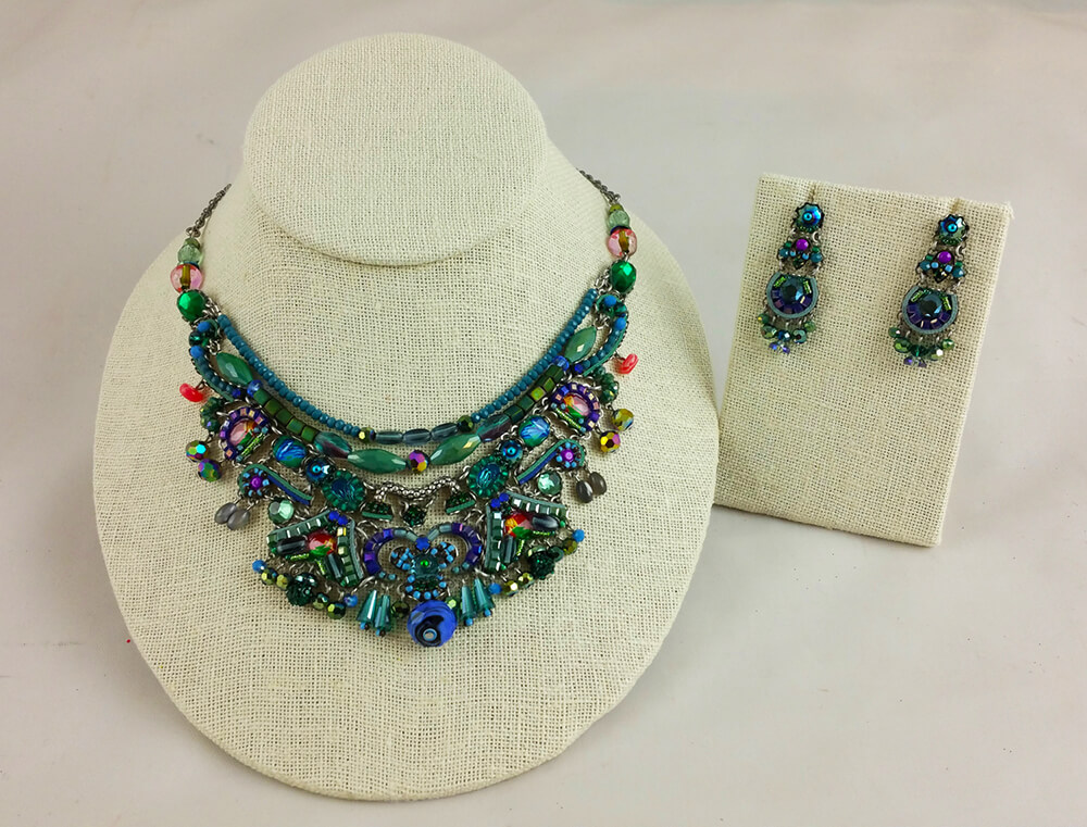 An elaborate, multicolored beaded necklace with matching earrings displayed on a white textured bust stand and earring holder. The jewelry features an intricate design with green, blue, purple, and red beads accented with metal elements.