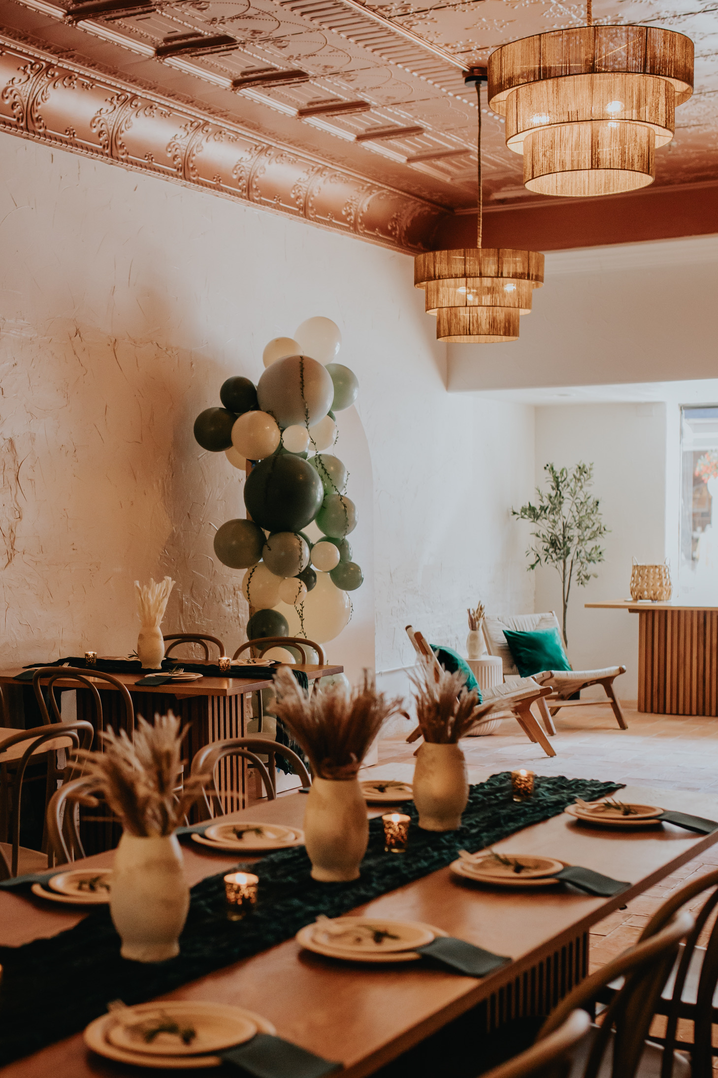A cozy, elegantly decorated room features a long wooden dining table set with white plates, light green napkins, and dried floral centerpieces. Wooden chairs surround the table. The ceiling has intricate designs and warm lighting, with a balloon decoration in the corner.