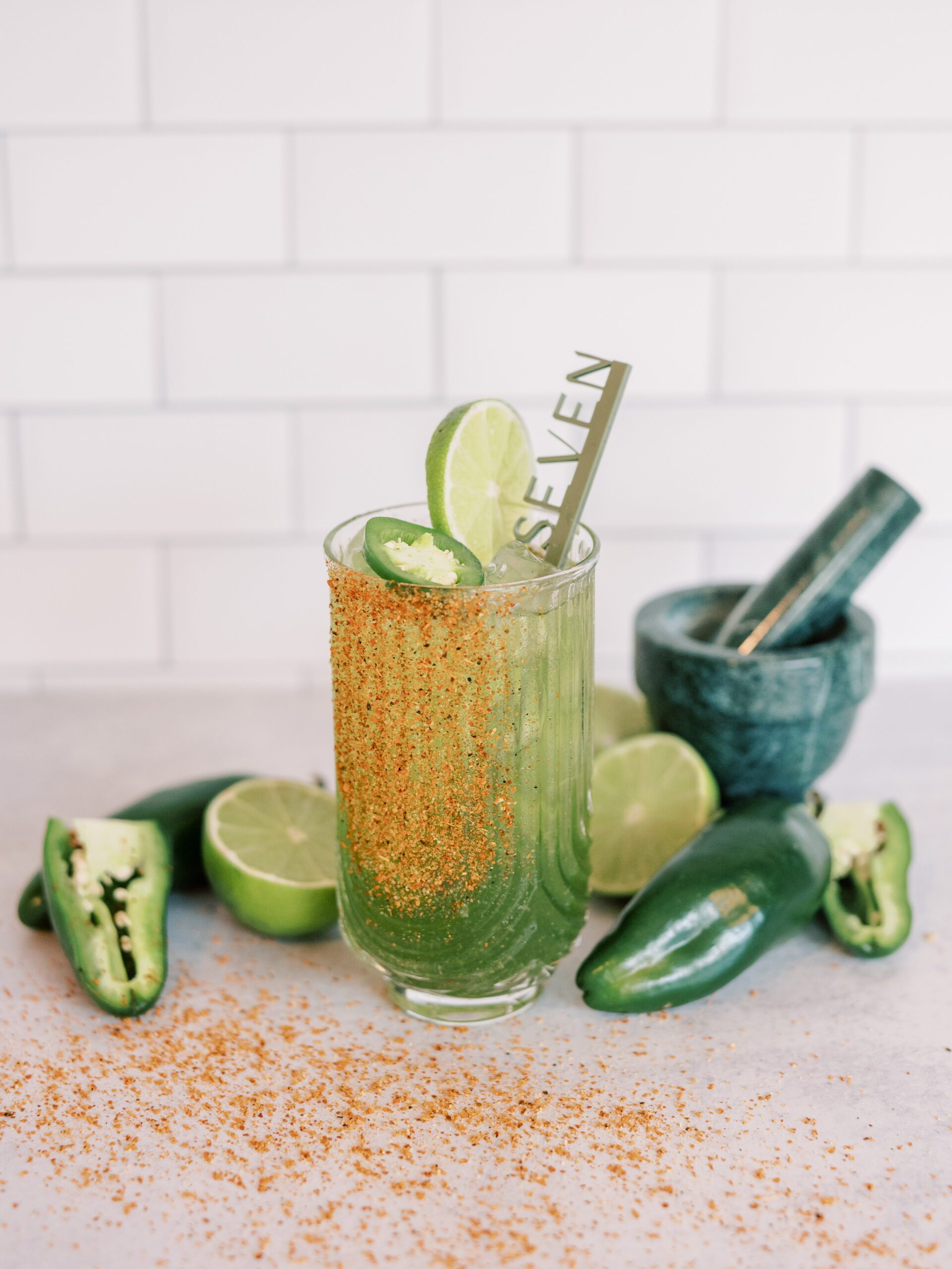 A tall glass filled with a green beverage garnished with lime slices and jalapeño, rimmed with chili powder. The drink is in front of sliced limes and jalapeños, with a green mortar and pestle set in the background. A straw labeled 