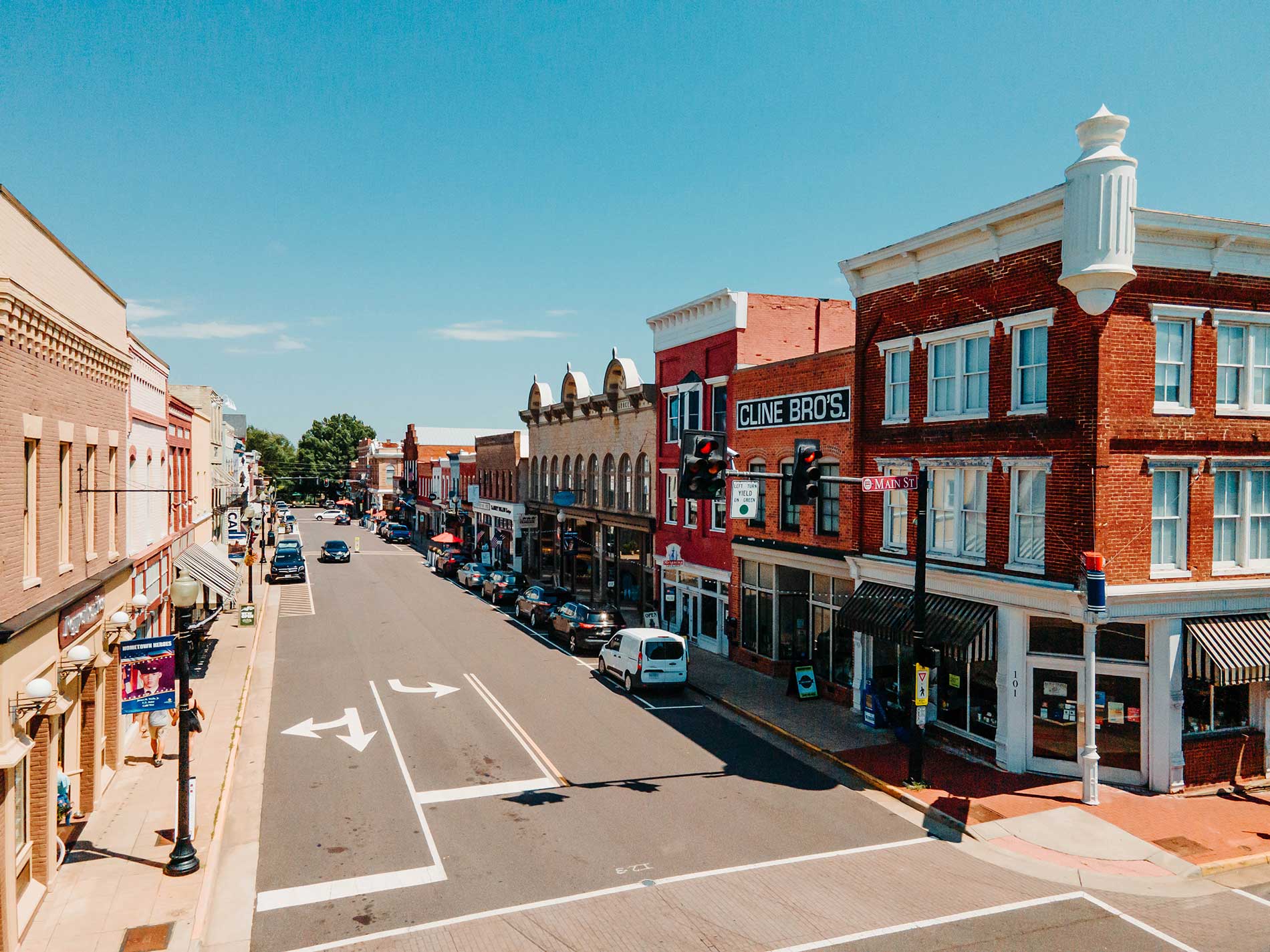 A bustling downtown street lined with historic brick buildings on a sunny day. Various shops and businesses, including 'Cline Bros,' are visible with their signage. Several cars are parked along the side of the street, and a few pedestrians are walking on the sidewalks.