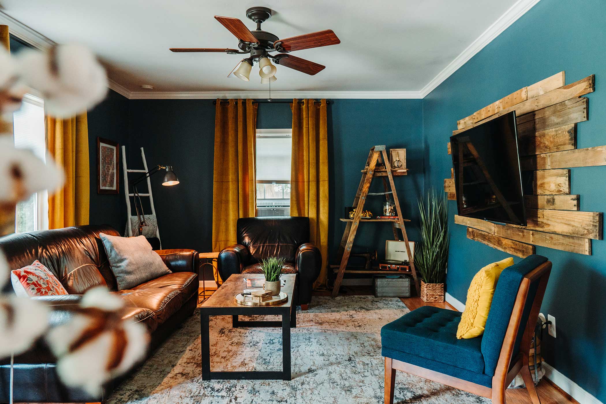 A cozy living room with blue walls and mustard yellow curtains. It features brown leather sofas, a blue accent chair, and a rustic wooden coffee table. A TV is mounted on a wooden pallet-styled wall. Various decorations and plants enhance the warm and inviting atmosphere.