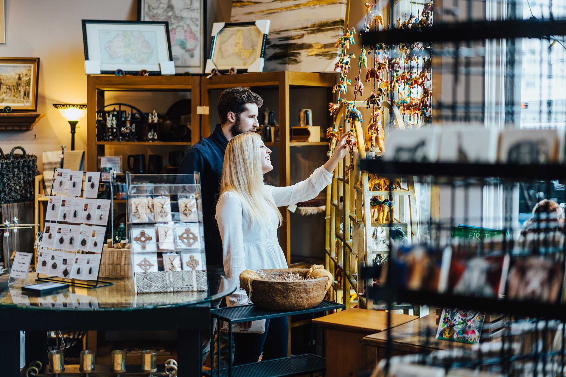 A man and woman are browsing items in a shop filled with various trinkets and artwork. The woman is reaching out to touch a display of hanging ornaments while the man stands next to her, observing. Shelves and tables around them hold cards, crafts, and framed prints.