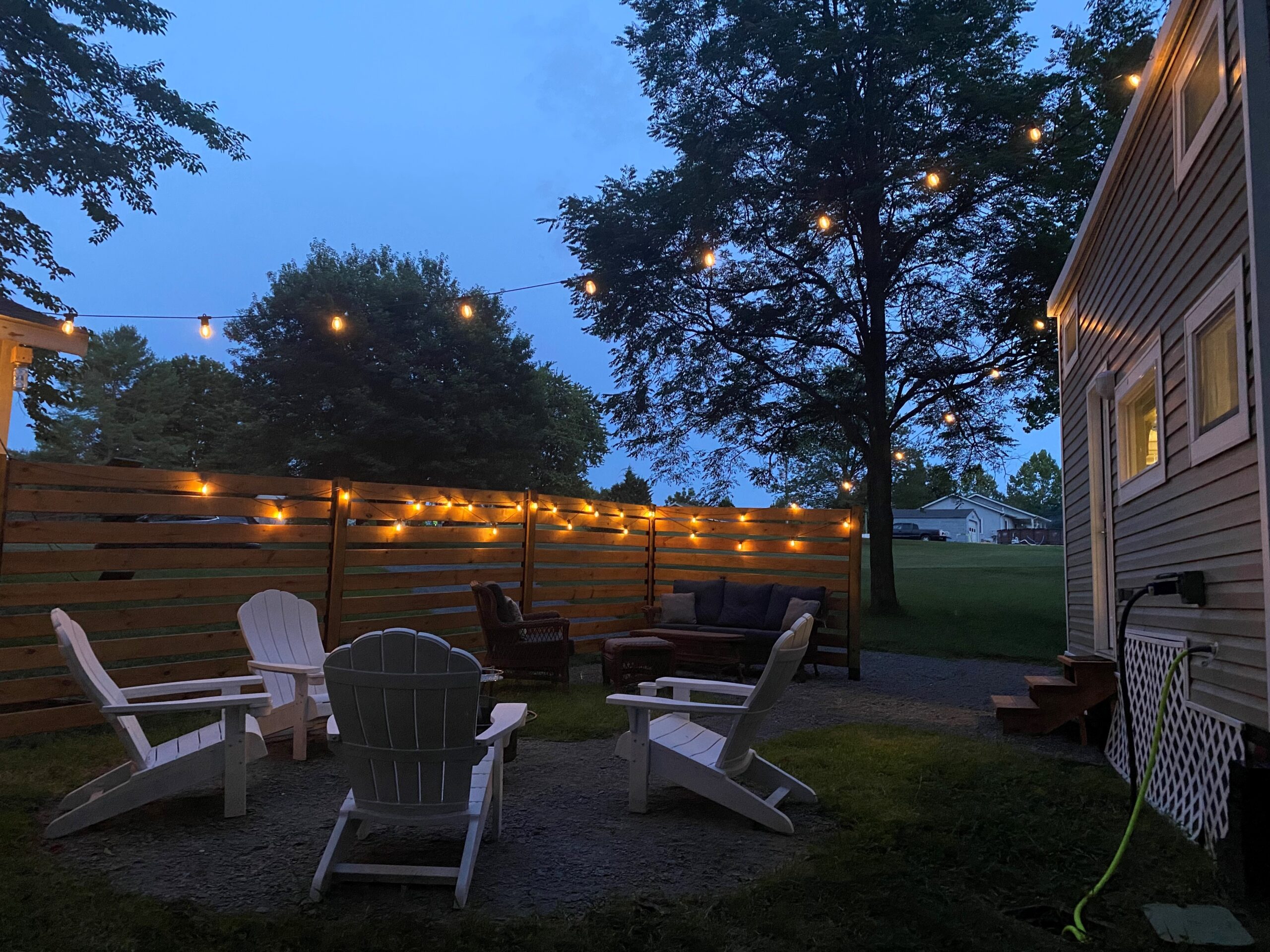 A cozy outdoor seating area with white Adirondack chairs surrounds a fire pit, perfect for your next Airbnb vacation rental. String lights hang above and along the wooden fence, illuminating the space. Nearby, a house with siding is partially visible, framed by trees and a grassy yard at dusk.