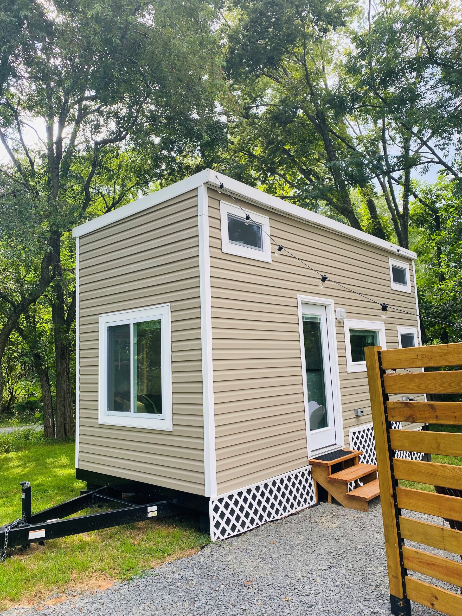 A tiny house with beige siding and white trim sits on a trailer in a wooded area. It features large windows, a small porch with steps leading up to it, and lattice skirting. The lush green trees and gravel path create an inviting scene, making it an ideal Airbnb vacation rental.