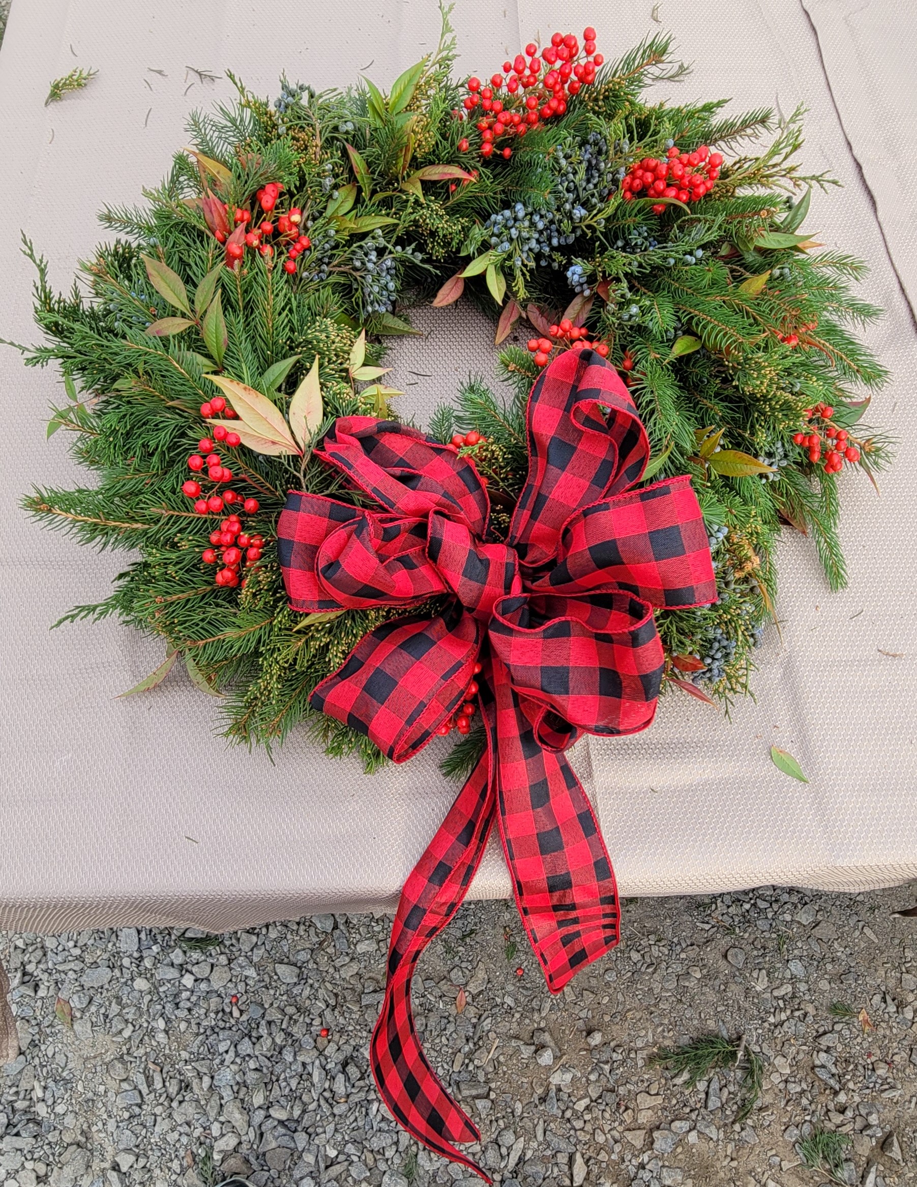 A festive holiday wreath made of green foliage and accented with red berries and small blue clusters. The wreath is adorned with a large red and black plaid bow with long ribbon tails, all set on a beige surface with gravel visible at the bottom.