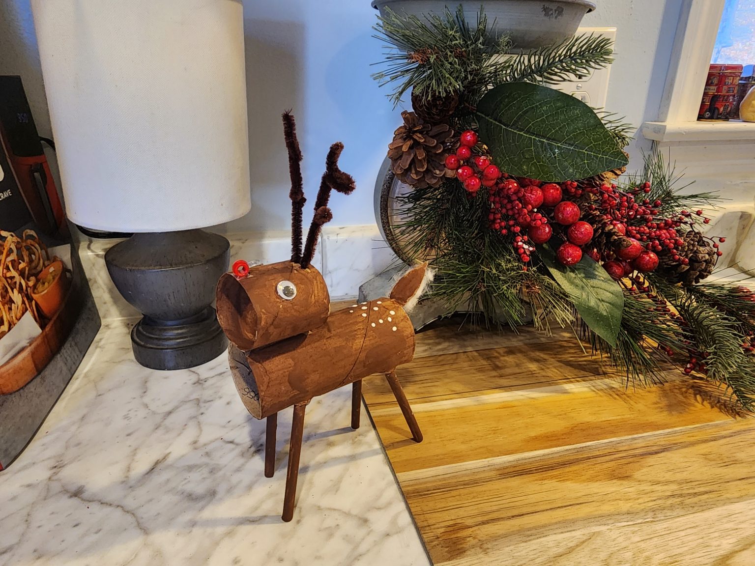 A handmade reindeer ornament with a red nose, googly eyes, and twig antlers stands on a marble surface. Next to it is a festive Christmas arrangement with pinecones, red berries, and green leaves. A lamp and decorative items in the background complete the cozy scene.