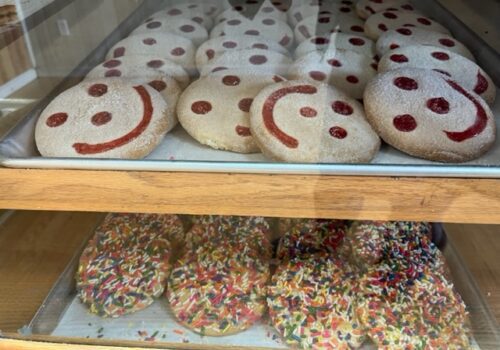 A display case with two trays of cookies. The top tray holds round cookies decorated with red smiley faces made of icing and small dots for eyes. The bottom tray has round cookies covered with colorful sprinkles. Both trays are on wooden shelves. Image