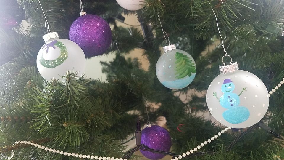A close-up of Christmas tree branches adorned with hand-painted ornaments. The ornaments feature designs like a wreath, a decorated tree, a snowman, and purple sparkly spheres. A pearl-like garland and black string lights are also visible on the tree.