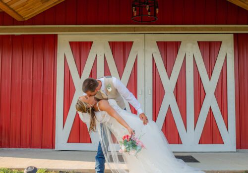A bride and groom share a kiss in front of a large, bright red barn. The groom, in a white shirt and light-colored vest, dips the bride, who is in a flowing white wedding gown with a long veil and holding a bouquet of pink flowers. Image