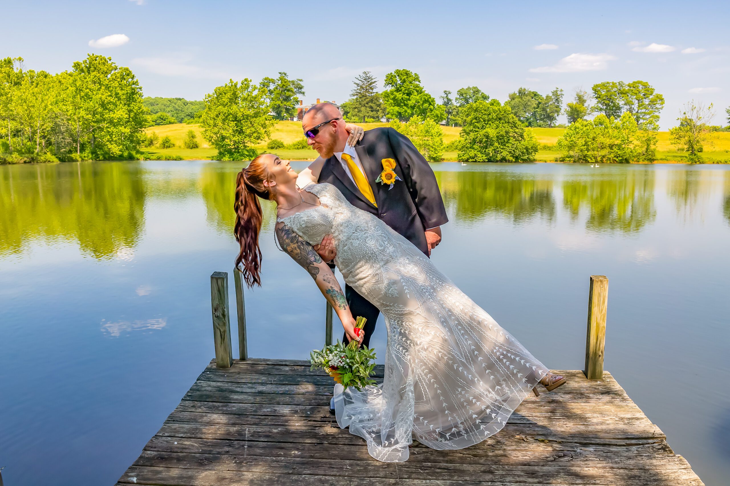 A tattooed bride in a white lace dress and a groom in a dark suit with a yellow tie share a playful moment on a wooden dock by a tranquil lake, surrounded by lush greenery and a clear blue sky. The groom leans back the bride as they both smile.