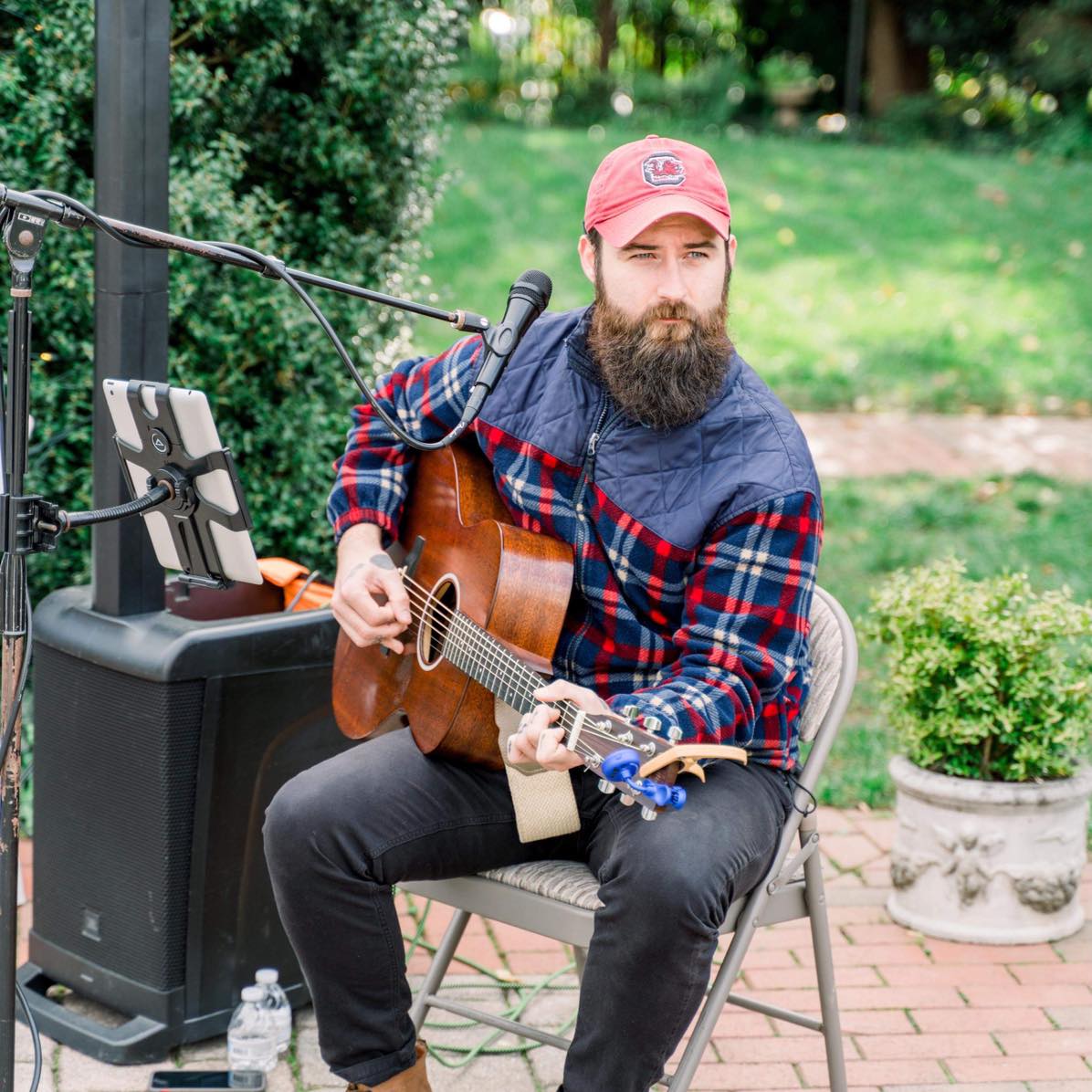 A bearded man in a red cap and plaid jacket sits outdoors playing an acoustic guitar. He is seated on a wooden chair, with a microphone stand, a water bottle, and some equipment around him. The background includes greenery and a garden.